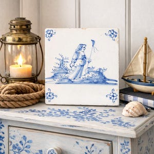 May include: A square, white ceramic tile with a blue and white illustration of a person holding a flag. The tile has floral corner designs. A lit candle in a brass lantern, a toy sailboat, and a seashell are also visible.