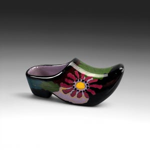 Puede incluir: Un zueco de cerámica con acabado negro brillante, que presenta un diseño floral pintado a mano con un centro amarillo brillante, pétalos magenta y hojas verdes. El interior del zapato está esmaltado en un suave color lavanda.