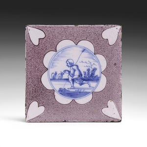 May include: Square ceramic tile with a blue and white illustration of a person with a spear, kneeling near two round objects. The illustration is within a white, flower-shaped border. The tile has a textured, mauve background with white heart-shaped accents.