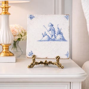 May include: A vintage, rectangular ceramic tile with a blue and white pastoral scene, displayed on an ornate gold-colored metal stand. The tile features a figure with a staff on a hill. The corners of the tile have blue floral accents.