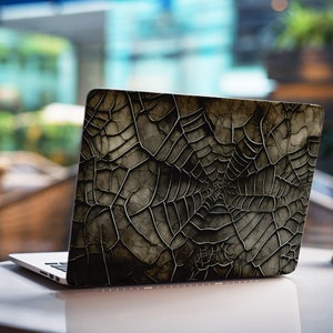 May include: A laptop computer with a black and grey spiderweb design on the back. The laptop is sitting on a white table.