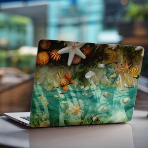 Puede incluir: Un ordenador portátil con una escena submarina colorida en la tapa. La escena presenta estrellas de mar, conchas y corales. El portátil está sobre una mesa blanca.