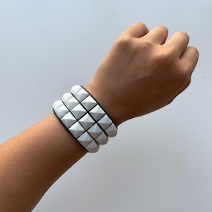 May include: A white studded bracelet with a black band is worn on a wrist. The bracelet features three rows of white pyramid-shaped studs. The background is a plain, light gray color.