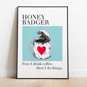 Może przedstawiać: Oprawiony wydruk artystyczny z ilustracją borsuka miodowego. Borsuk znajduje się w kubku do kawy z czerwonym sercem. Tekst brzmi "HONEY BADGER" nad obrazem i "First I drink coffee, then I do things" poniżej.