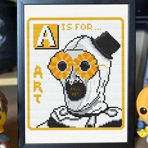 May include: A cross-stitch pattern with a white background and black frame. The pattern is a letter A with the words "A is for... ART" and a character with a white mask and yellow sunglasses.