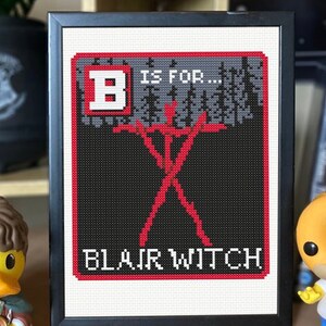 May include: A black framed cross-stitch with a red letter B, the words "B is for... Blair Witch", and a red stick figure in a forest.