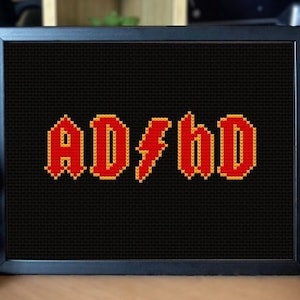 Peut inclure: Une broderie au point de croix encadrée en noir avec le texte "AD#HD" en lettres rouges et orange sur un fond noir. La broderie est posée sur une surface en bois avec deux figurines de canards en caoutchouc de chaque côté.