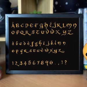 May include: A black framed cross-stitch with a gold alphabet, numbers, and punctuation marks. The alphabet is in a medieval font. The cross-stitch is on a shelf with other items.