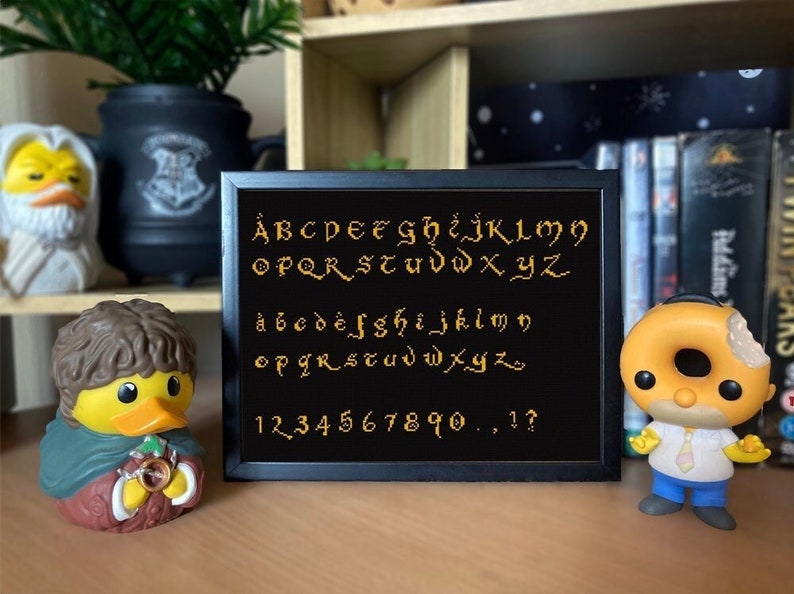 May include: A black framed cross-stitch with a gold alphabet, numbers, and punctuation marks. The alphabet is in a medieval font. The cross-stitch is on a shelf with other items.