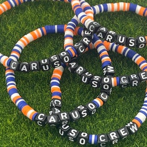 Puede incluir: Varios brazaletes con la palabra "CARUSO CREW" escrita en letras blancas sobre cuentas cuadradas negras. Los brazaletes están hechos de cuentas planas naranjas, blancas y azules. Los brazaletes están sobre una superficie verde.