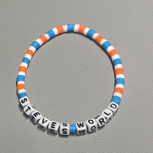 Puede incluir: Un brazalete de cuentas blancas con cuentas azules y naranjas alternas. Las cuentas deletrean "STEVE'S WORLD" en letras negras.
