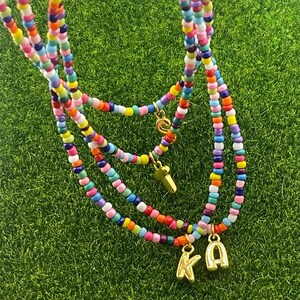 May include: A multi-strand beaded necklace with colorful beads in various sizes. The necklace has gold-colored charms and initial pendants. The background is green.