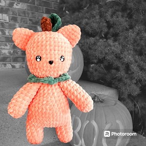 May include: A crocheted plush toy shaped like a pumpkin with a green and brown stem. The toy is peach colored and has a friendly face with black eyes and a black nose. The toy is sitting on a table with a pumpkin behind it.