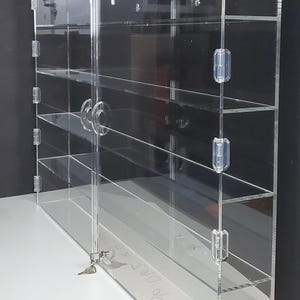 May include: A clear acrylic display case with multiple shelves and a hinged door. The case has a lock and key, and is designed to showcase collectibles or other items. The case is on a white surface.