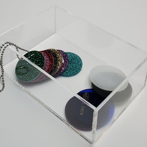 May include: A clear acrylic box contains several round, glittery items in various colors, including green, pink, gold, and teal. A silver ball chain is attached to one of the items. Additional items include a white oval and a dark blue disc.