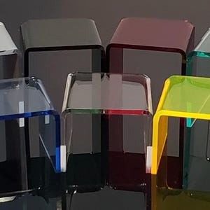 May include: A collection of small, square acrylic display stands in various colors, including clear, red, blue, green, yellow, and white. The stands have a modern, minimalist design, perfect for showcasing small items.