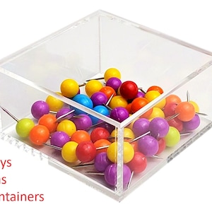 May include: A clear acrylic square container filled with colorful push pins. The push pins are various colors including yellow, orange, red, purple, and blue. The text "Trays, Bins, Containers" is visible in the lower left corner.