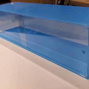 May include: A light blue acrylic display case with a clear glass front and a shelf inside. The case has a small hole on the side.