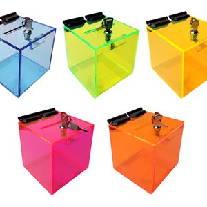 May include: Five transparent acrylic donation boxes in blue, green, yellow, pink, and orange. Each box has a slot on top, a black hinge, and a silver lock with a key. The boxes are cube-shaped.