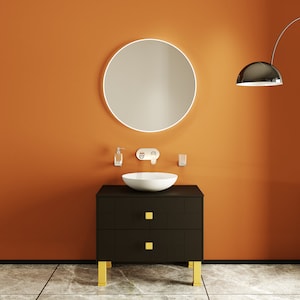 May include: A black bathroom vanity with two drawers and gold legs. The vanity has a white ceramic sink and a round mirror with a white frame. The wall is painted a bright orange.