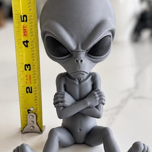 May include: A gray alien figurine with a large head and black eyes, sitting with arms crossed. A yellow measuring tape is positioned next to the figure, showing a height of approximately 6.5 inches from the base to the top of the head.