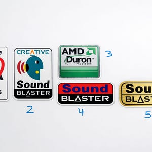 Peut inclure: Plusieurs autocollants rectangulaires avec des logos, dont "3dfx Voodoo Graphics", "Creative Sound Blaster", et "AMD Duron Processor". Deux designs "Sound Blaster" sont également présents, un rouge et un doré.
