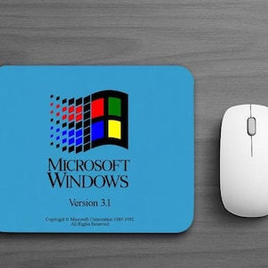 May include: A rectangular light blue mouse pad featuring the classic Microsoft Windows Version 3.1 logo with red, green, blue, and yellow squares, accompanied by a white wireless computer mouse positioned to the right of the pad.