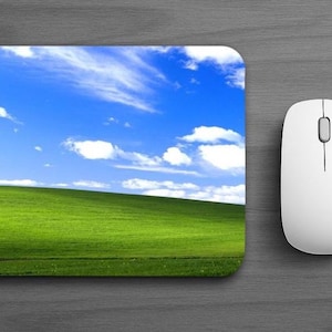 May include: A rectangular mouse pad featuring a vibrant landscape print of a bright green grassy hill under a clear blue sky with scattered white clouds, positioned next to a sleek white computer mouse with a central scroll wheel.
