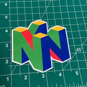 Puede incluir: Una pegatina con el logotipo de Nintendo 64. El logotipo es una 'N' tridimensional en verde, azul, rojo y amarillo, con un borde blanco. La pegatina está sobre una esterilla de corte verde.