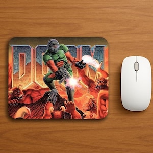 Puede incluir: Alfombrilla de ratón rectangular con una vibrante escena de acción del videojuego "Doom". La obra de arte representa a un personaje vestido de verde luchando contra criaturas demoníacas, con el título del juego en un lugar destacado. Un ratón de ordenador blanco está a la derecha.