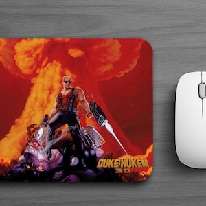 May include: A rectangular mouse pad featuring a graphic of the character Duke Nukem standing before a large orange and red explosion. The text 3D Realms appears in the top left corner, and the text Duke Nukem 3D is printed in yellow at the bottom.