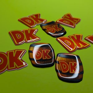 May include: A collection of DK stickers and barrel-shaped stickers on a green background. The DK stickers have red letters with a yellow outline. The barrel stickers are brown with the DK logo.