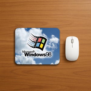 May include: A rectangular mousepad featuring the Windows 98 logo against a blue sky with white clouds. The mousepad is next to a white wireless mouse on a wooden surface.