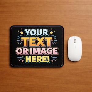May include: A black rectangular mousepad with the text "YOUR TEXT OR IMAGE HERE!" in colorful lettering. A white wireless mouse is positioned to the right of the mousepad. The background is a wooden surface.