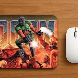 May include: A rectangular mouse pad featuring the DOOM video game cover art. A green-armored soldier fires a weapon at red, horned demonic creatures against a backdrop of the large metallic DOOM logo. The scene is filled with fiery orange tones.