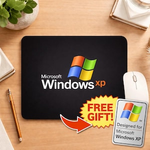 May include: Black mousepad featuring the Microsoft Windows XP logo in white, orange, blue, and green. Includes a "FREE GIFT!" sticker and a white mouse. The mousepad is rectangular and sits on a wooden desk.