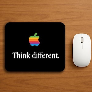May include: Black rectangular mousepad featuring the Apple logo in rainbow colours and the words "Think different." A white wireless computer mouse is to the right. The items are on a wood grain surface.