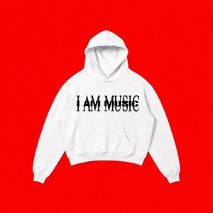 May include: A white hooded sweatshirt featuring a front kangaroo pocket and long sleeves. The chest displays the text I AM MUSIC printed in a bold black serif font, with a secondary overlapping black text effect creating a layered visual.