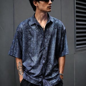 Peut inclure: Une chemise à manches courtes boutonnée avec un motif peau de serpent bleu foncé et noir. La chemise présente un col classique, une patte de boutonnage sur le devant et une coupe décontractée. Le tissu a un léger éclat, soulignant la texture détaillée des écailles reptiliennes sur tout le vêtement.