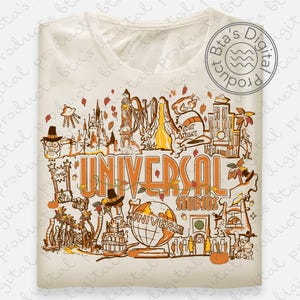 May include: Cream-colored t-shirt featuring a detailed illustration of Universal Studios attractions in shades of orange and brown. The text "UNIVERSAL STUDIOS" is prominently displayed. The design includes a globe, roller coasters, and other park elements.