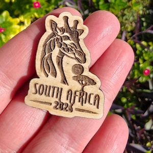 May include: A wooden souvenir featuring a giraffe design and the text "SOUTH AFRICA 2023". The giraffe is intricately carved, and the text is in a bold, readable font. The souvenir is a light brown color.