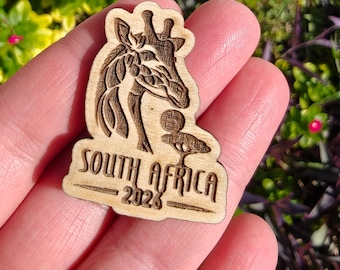 May include: A wooden souvenir featuring a giraffe design and the text "SOUTH AFRICA 2023". The giraffe is intricately carved, and the text is in a bold, readable font. The souvenir is a light brown color.