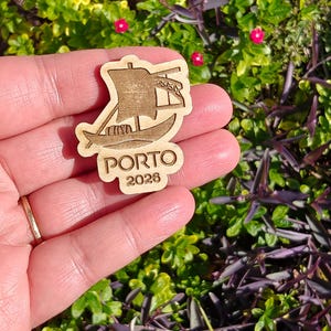 May include: A wooden cutout of a boat with a sail, featuring the word "PORTO" and the year "2026". The cutout is held in a hand against a backdrop of green foliage and small pink flowers. The boat design is etched into the wood.