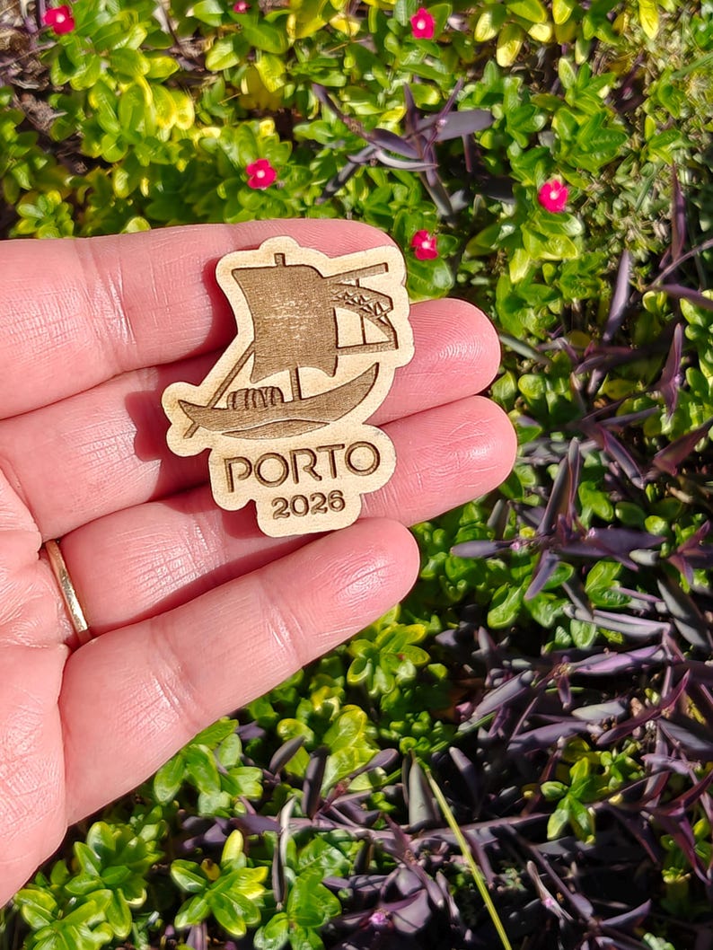 May include: A wooden cutout of a boat with a sail, featuring the word "PORTO" and the year "2026". The cutout is held in a hand against a backdrop of green foliage and small pink flowers. The boat design is etched into the wood.