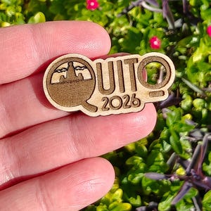 May include: A wooden pin featuring the word "QUITO" and the year "2026". The pin has a cutout design of a landscape within the letter "Q". The pin is made of light brown wood.