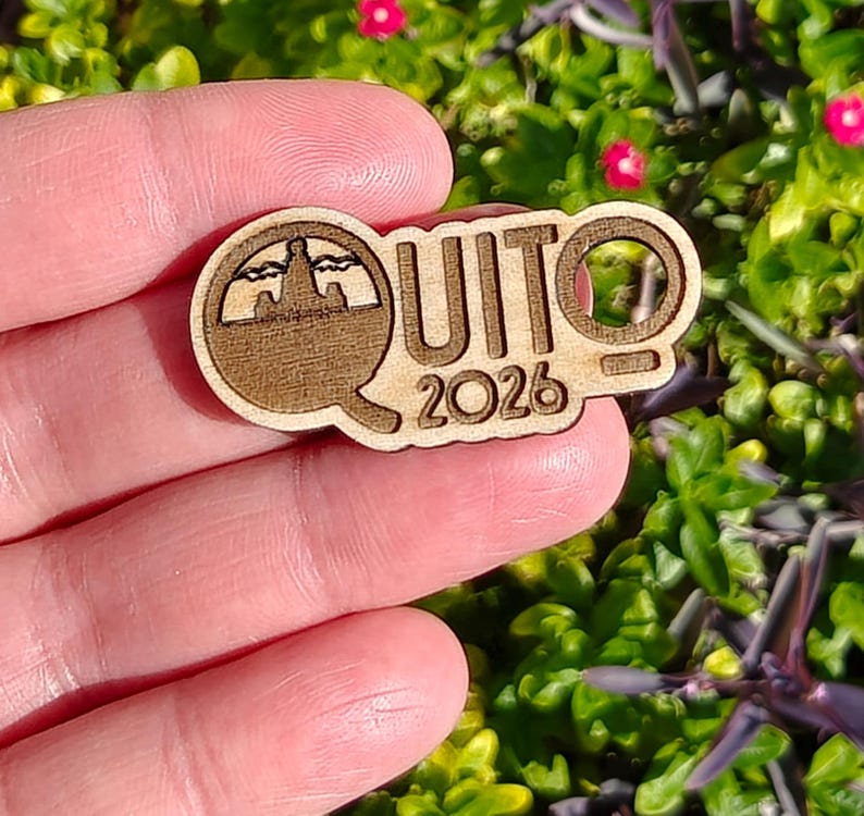 May include: A wooden pin featuring the word "QUITO" and the year "2026". The pin has a cutout design of a landscape within the letter "Q". The pin is made of light brown wood.