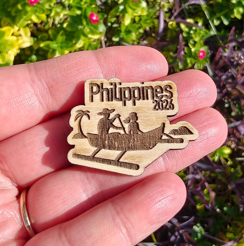 May include: Wooden Philippines souvenir with a boat scene and the text "Philippines 2026". The design features a palm tree and two figures in a boat. The souvenir is made of light-colored wood.