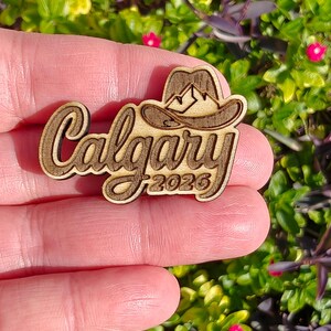 May include: Wooden Calgary souvenir with a cowboy hat and mountain design. The word "Calgary" is in a cursive font, with "2026" below. The item is held in a hand against a background of green and purple foliage.
