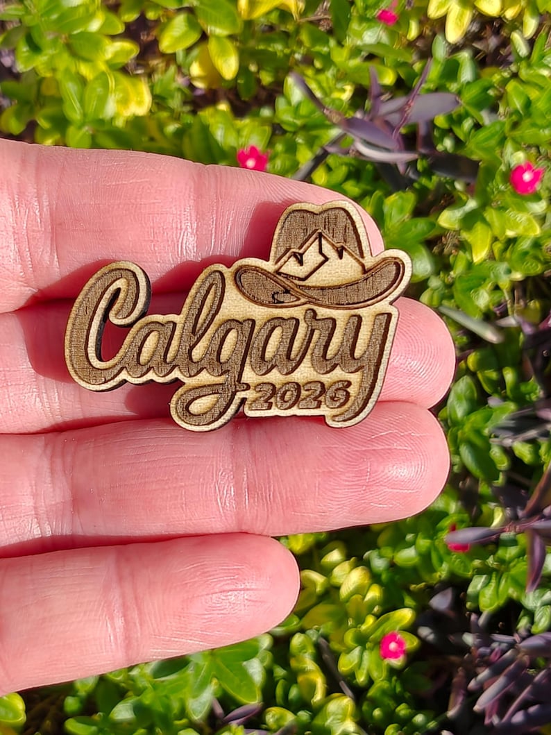 May include: Wooden Calgary souvenir with a cowboy hat and mountain design. The word "Calgary" is in a cursive font, with "2026" below. The item is held in a hand against a background of green and purple foliage.