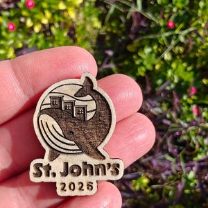 May include: Wooden emblem featuring a whale with buildings in its body, and the text "St. John's 2026". The emblem is made of light-colored wood with a darker, engraved design. The whale's tail curves around the buildings.